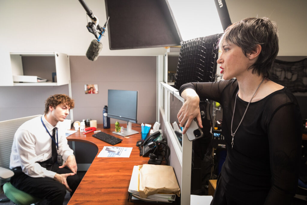 From the set of Sick Days, Jose and Chelsea stand in an office separated by a cubicle wall