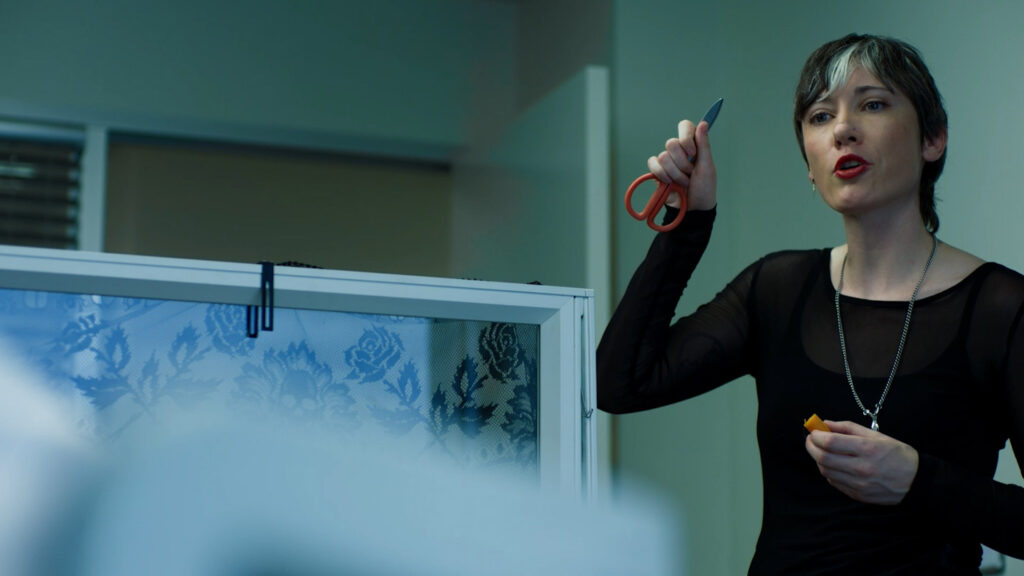 From Sick Days, a woman holds up a pair of scissors in a cubicle